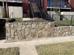 Completed natural stone retaining wall along a commercial property sidewalk with metal stair railing and brick building in Arlington TX
