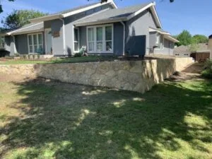 New Granbury stone retaining wall running across front yard of gray DFW home, replacing a failing structure