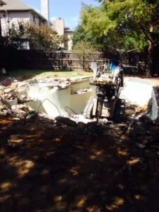 Excavator breaking apart inground pool shell during residential pool removal in Fort Worth TX backyard, debris and rubble visible