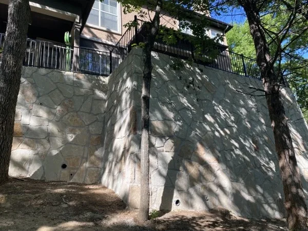 Tall Milsap stone retaining wall supporting elevated condo deck with black iron railing, shaded by mature trees in Arlington TX