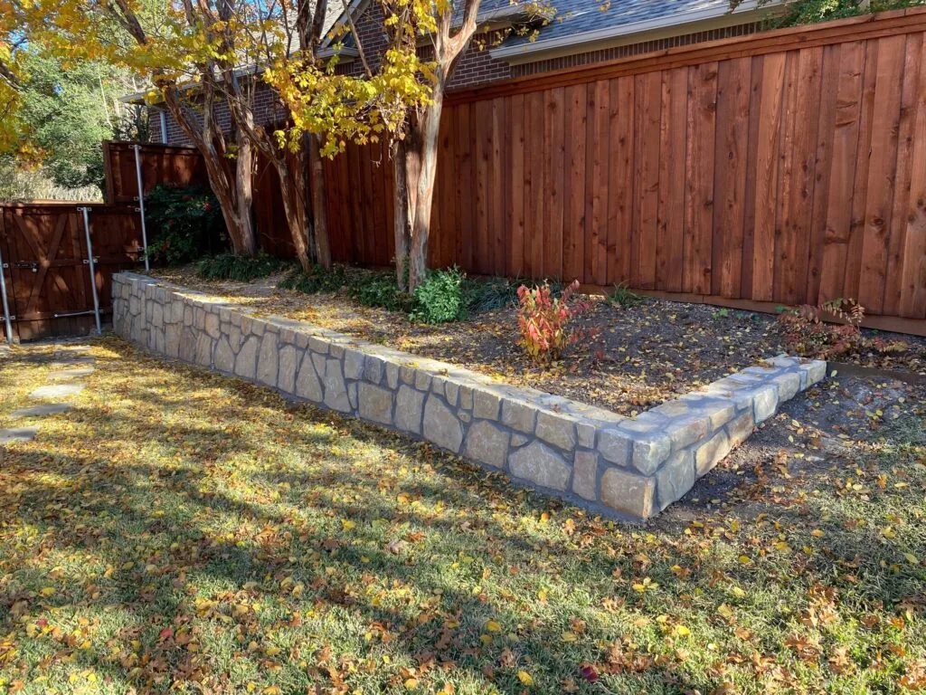 L-shaped Milsap stone retaining wall forming a raised planting bed in a Carrollton, TX backyard beside a wood privacy fence