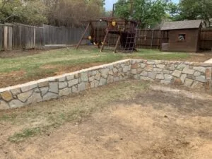 New Milsap stone retaining wall replacing old crosstie wall in a Bedford TX backyard with playground set behind