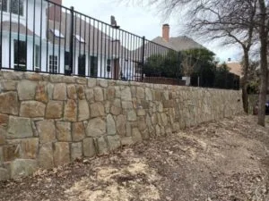 Rebuilt Milsap stone retaining wall with wrought-iron fence atop, replacing failed cross-tie wall at Coppell TX residence