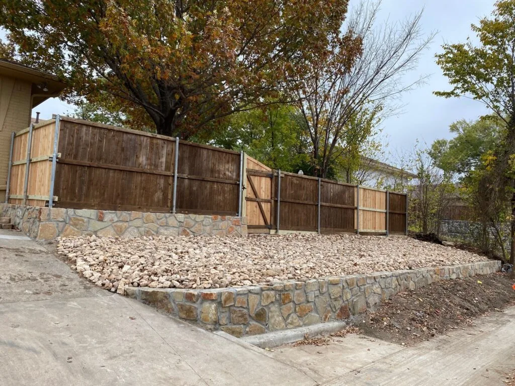 Natural stone retaining wall with gravel infill supporting wood privacy fence along residential driveway in Carrollton TX