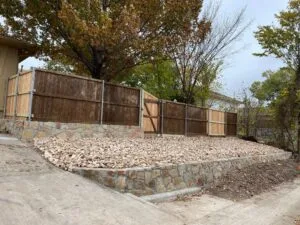 Natural stone retaining wall with gravel infill supporting wood privacy fence along residential driveway in Carrollton TX