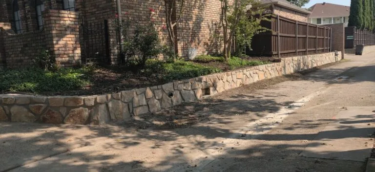 Natural stone retaining wall with drainage weep holes along a brick home's driveway, holding landscaped garden bed in Fort Worth TX