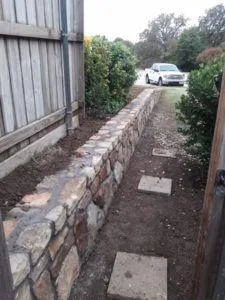 Newly built natural stone retaining wall along a residential fence line in Lewisville TX, with fresh backfill and truck in driveway