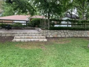 Natural stone retaining wall with integrated steps replacing old cross-tie wall in Denton TX backyard, creating level patio terrace