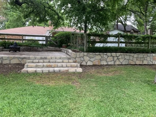 Natural stone retaining wall with integrated steps replacing old cross-tie wall in Denton TX backyard, creating level patio terrace
