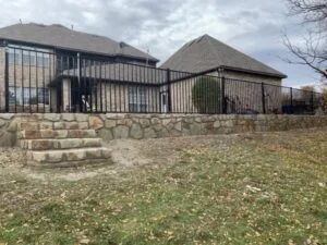 Natural stone retaining wall with integrated steps leveling a sloped backyard behind a brick home, black iron fence above, Frisco TX