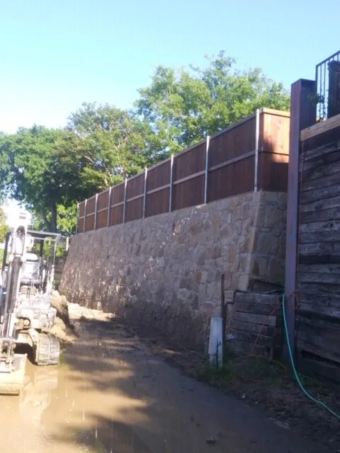 Large stone retaining wall under construction with wood fence on top and mini excavator on site in Fort Worth TX