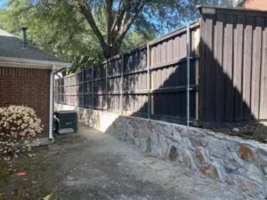 Completed natural stone retaining wall replacing old crosstie wall along dark wood privacy fence beside brick home in Carrollton TX