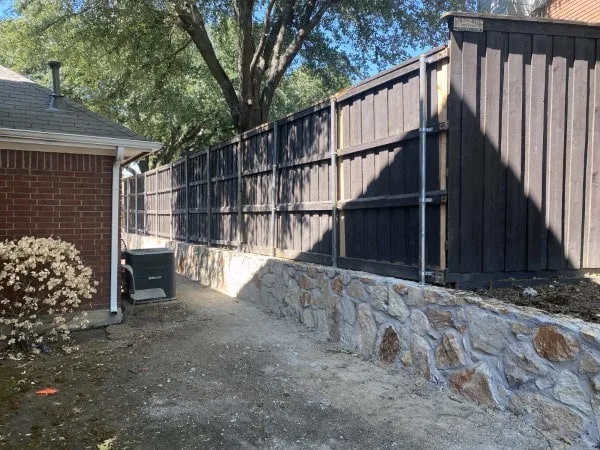 Completed natural stone retaining wall replacing old crosstie wall along dark wood privacy fence beside brick home in Carrollton TX