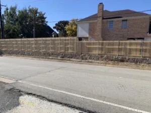 Stone retaining wall supporting a wood privacy fence along a residential road in Trophy Club TX, holding back elevated yard near brick home