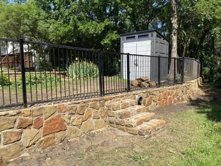 Natural Milsap stone retaining wall with built-in steps and black iron fence topping a raised backyard in Fort Worth TX