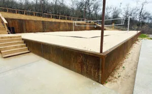 Weathered steel retaining walls contain a sand volleyball court with wood staircase and upper timber wall in Fort Worth TX