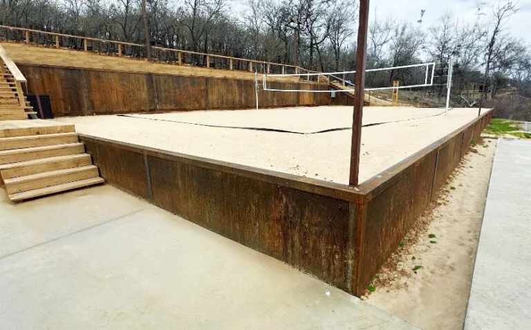 Weathered steel retaining walls contain a sand volleyball court with wood staircase and upper timber wall in Fort Worth TX