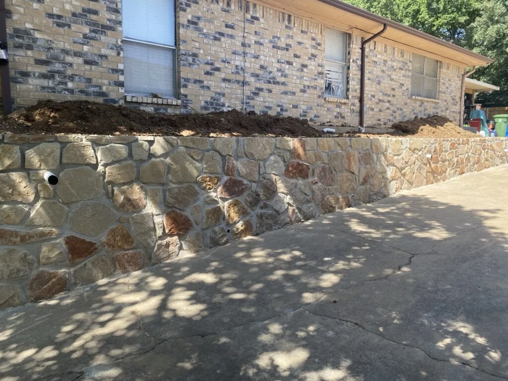 New Milsap stone retaining wall along brick home driveway in North Richland Hills, replacing old cinder block wall, with drainage weep holes and fresh backfill soil visible