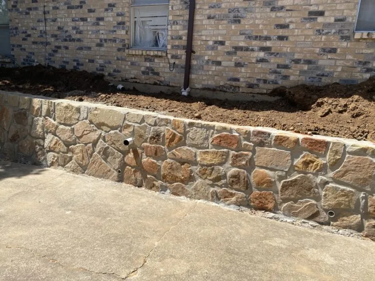 New Milsap stone retaining wall with weep holes along concrete driveway, replacing old cinder block wall beside brick home in North Richland Hills TX