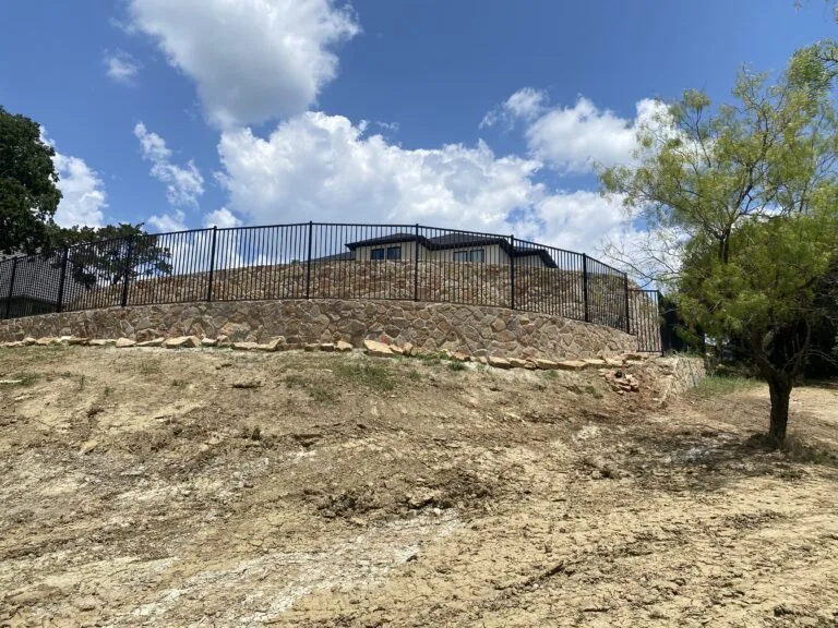 New natural stone retaining wall with black iron fence topping a graded slope behind a Fort Worth-area home, stabilizing expansive clay soil