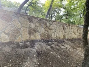 New Milsap stone retaining wall with drainage weep holes replacing old crosstie wall on wooded residential slope in Bedford TX