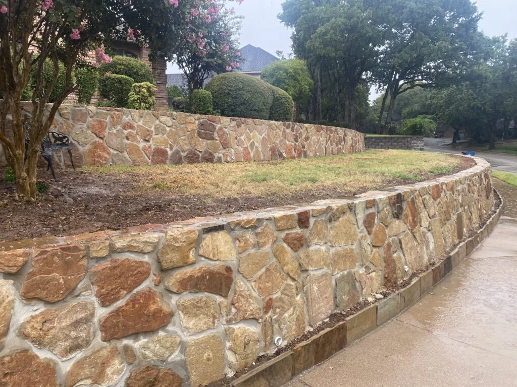 Curved two-tier natural stone retaining wall along a residential driveway in Grapevine TX, holding a terraced yard with crepe myrtles and brick home above