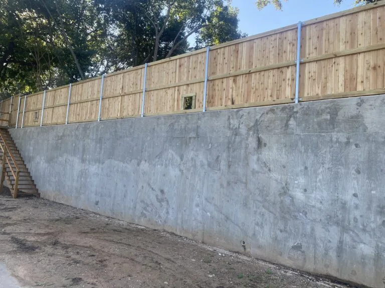 Tall poured concrete retaining wall with cedar fence on top and metal posts, holding back a sloped yard with exterior staircase in Fort Worth TX