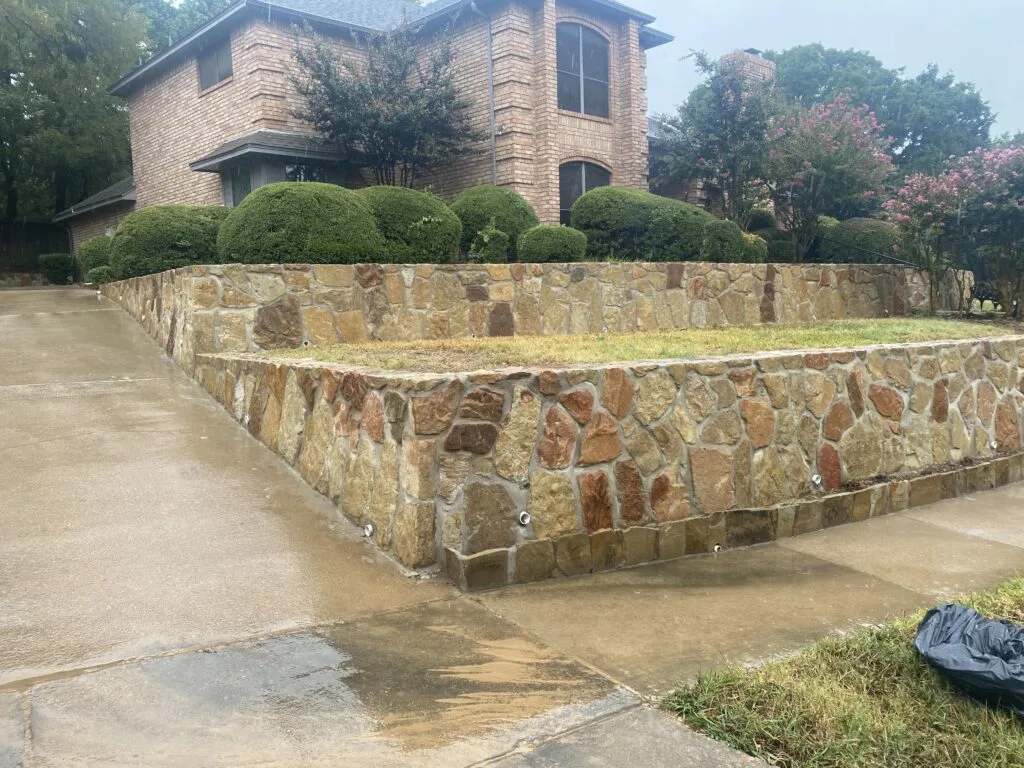 Two-tier natural stone retaining wall along a concrete driveway at a brick home in Grapevine TX, with drainage weep holes visible