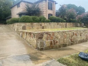 Two-tier natural stone retaining wall along a concrete driveway at a brick home in Grapevine TX, with drainage weep holes visible