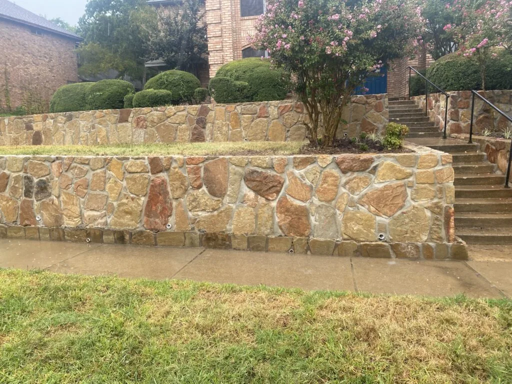 Two-tier natural stone retaining wall with drainage weep holes along a sloped front yard in Grapevine TX, with concrete steps and brick home above