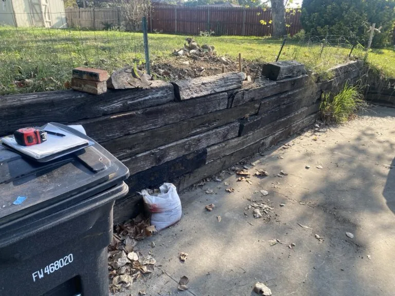 Failing railroad tie retaining wall with rotting timbers, soil erosion on top, and estimate notebook beside driveway in Fort Worth TX