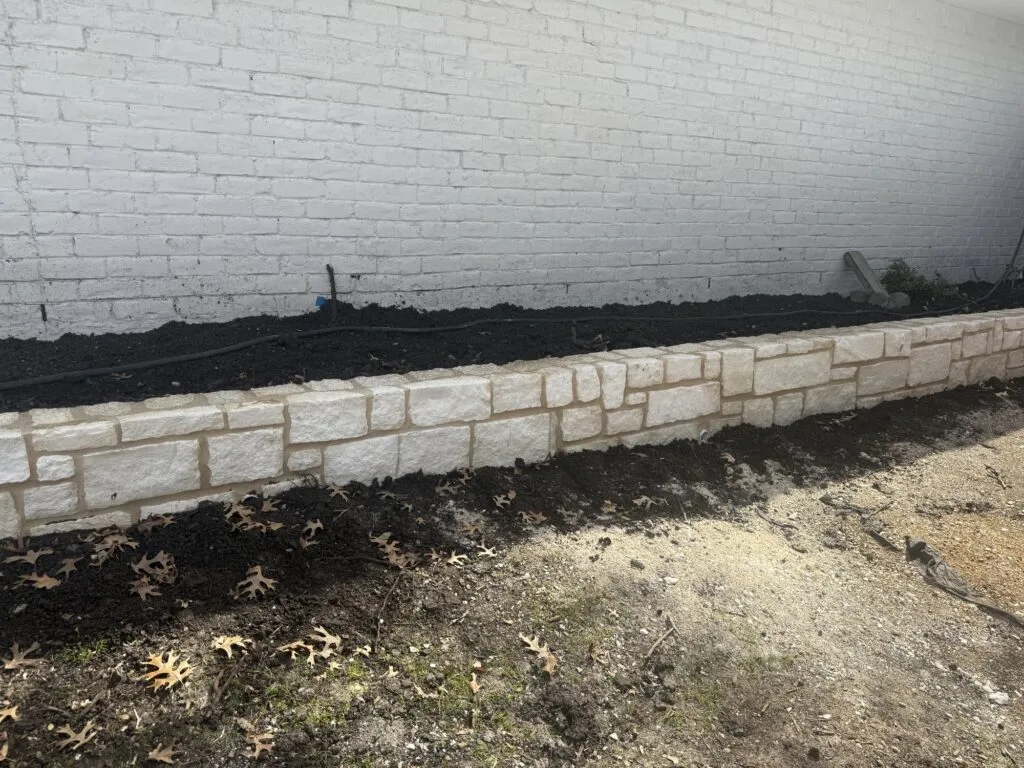 Newly built chopped Austin stone retaining wall along white brick building in Dallas, holding fresh dark soil bed