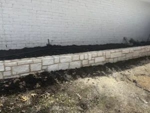 Newly built chopped Austin stone retaining wall along white brick building in Dallas, holding fresh dark soil bed