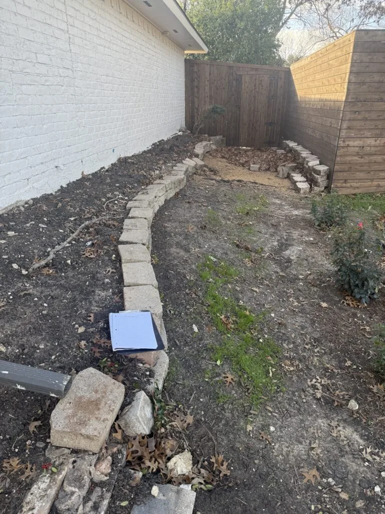 Failed low stone block retaining wall along brick home side yard during assessment, clipboard notes visible, Dallas TX