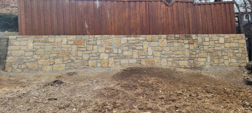 Newly built Granbury stone retaining wall with drainage weep holes along a wood fence line in Highland Village TX, fresh backfill grading visible