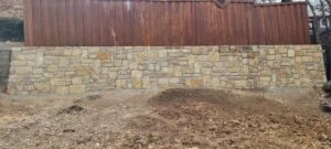 Newly built Granbury stone retaining wall with drainage weep holes along a wood fence line in Highland Village TX, fresh backfill grading visible