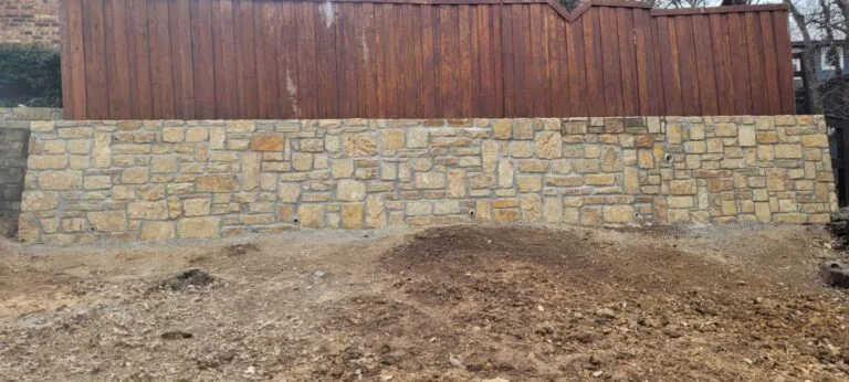 Newly built Granbury stone retaining wall with drainage weep holes along a wood fence line in Highland Village TX, fresh backfill grading visible