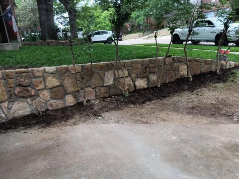 Natural stone retaining wall along a residential driveway in Grapevine TX, holding a raised planting bed with young trees