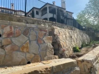 Large natural stone retaining wall with mixed tan and rust flagstone supporting elevated yard and iron fence behind Dallas home