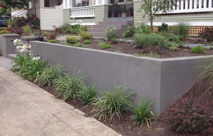 Smooth poured concrete retaining wall along a residential front yard, holding tiered planting beds beside a sidewalk and home entry steps