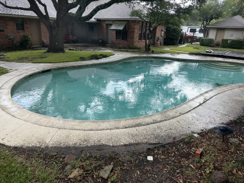 Aging kidney-shaped residential pool with cracked concrete coping filling with rainwater in a DFW backyard before pool removal demolition
