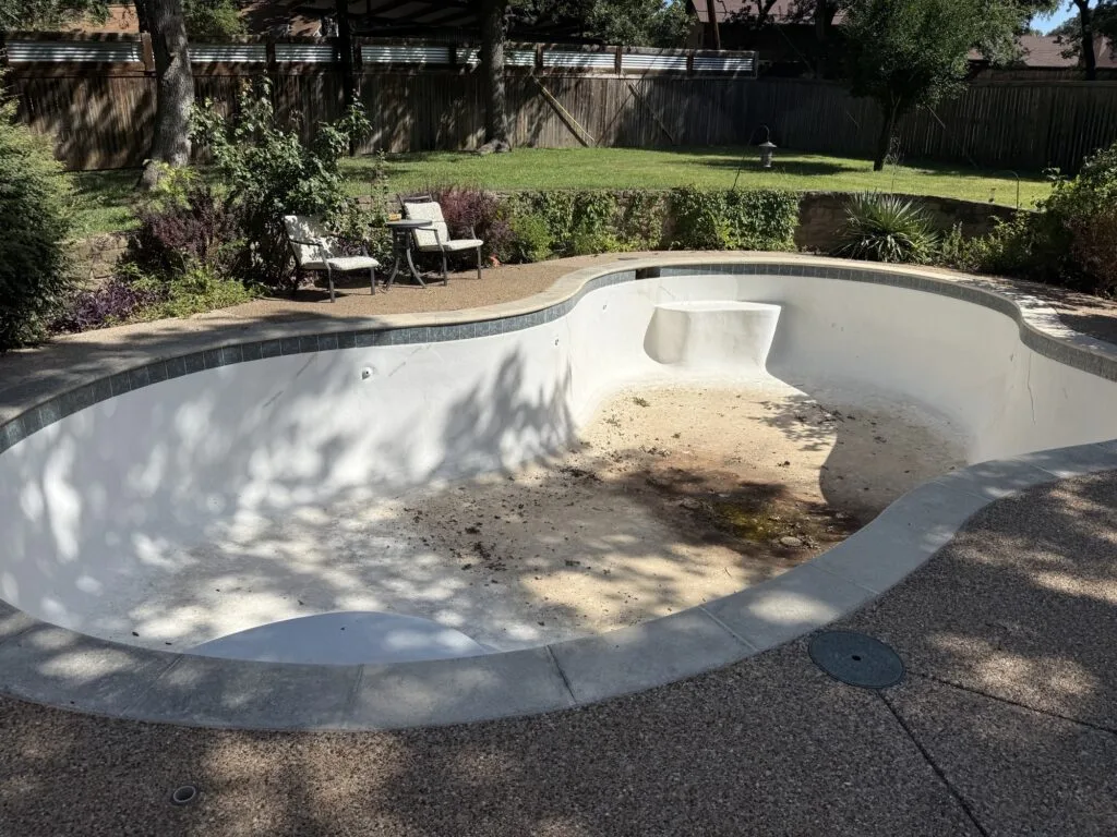 Drained kidney-shaped inground pool with cracked plaster and debris on floor, ready for demolition in Plano TX backyard