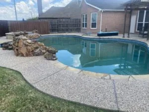 Freeform backyard pool with natural rock waterfall feature and aggregate concrete surround at a Fort Worth home before removal or wall work