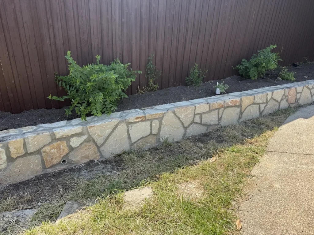 New mortared natural stone border wall with weep holes and a replanted bed along a fence line in Carrollton, TX