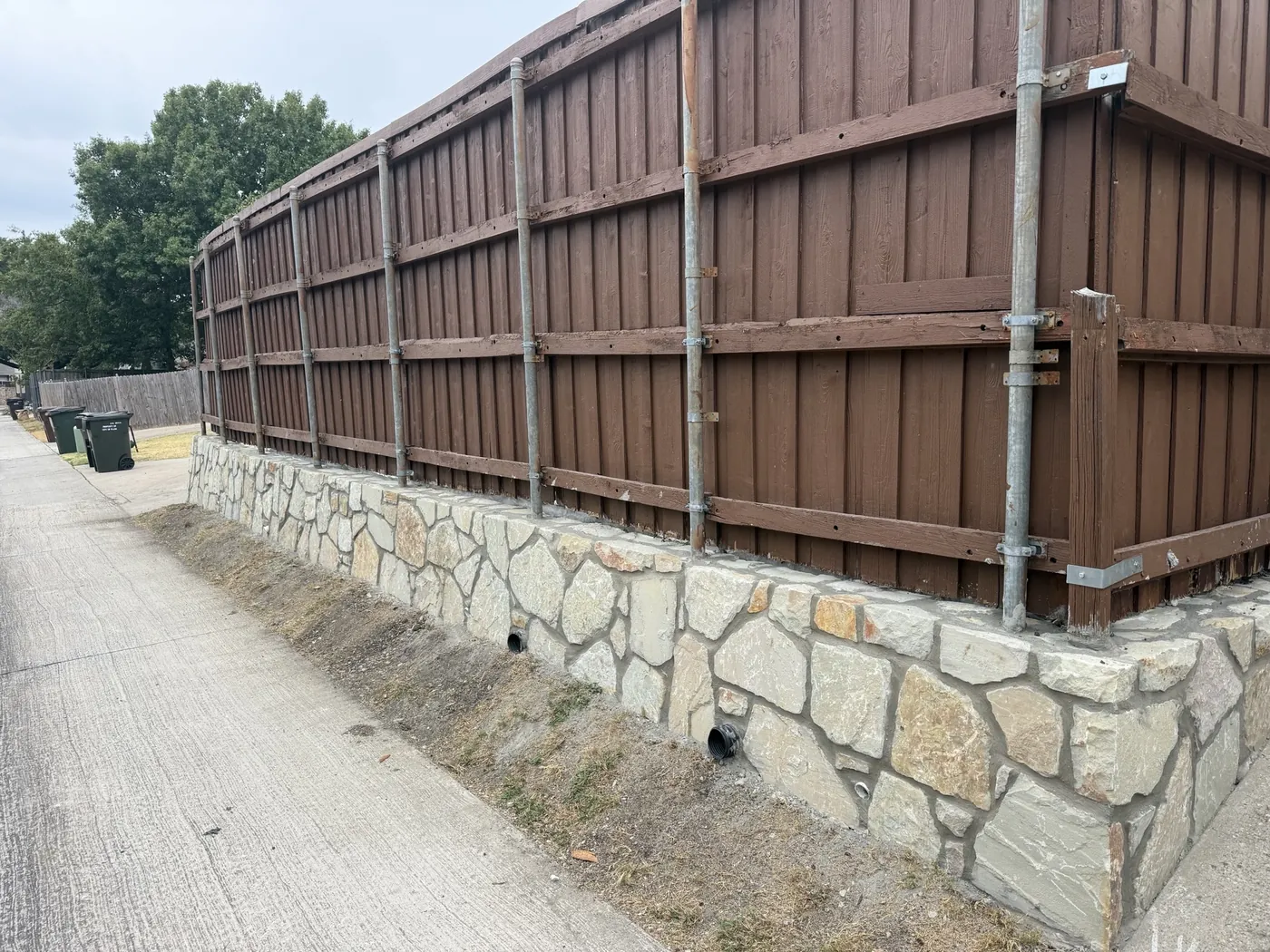 New mortared Milsap stone retaining wall with drain outlets under an existing fence along a Plano, TX alley